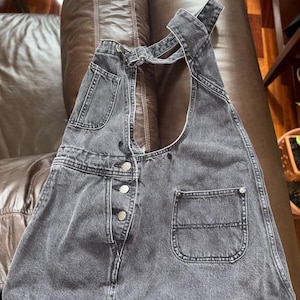 May include: A dark gray denim overall skirt with a single button closure and a large pocket on the right side. The straps are adjustable and cross in the back.