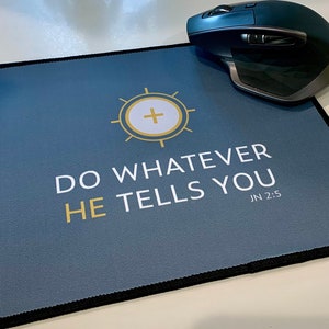 Pode incluir: Um tapete de rato retangular com fundo azul escuro e borda preta. O tapete apresenta um design dourado e branco com o texto "DO WHATEVER HE TELLS YOU" e "JN 2:5". Um rato sem fios cinza escuro está em cima.