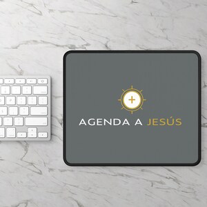 Op de afbeelding: Een grijze muismat met een zwarte rand. De muismat heeft een gouden zon met een kruis in het midden en de tekst "AGENDA A JESÚS" in wit.