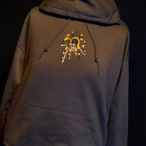 May include: A dark brown hooded sweatshirt with a black and orange embroidered spider on the front.