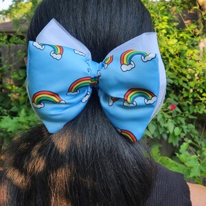 Può includere: Un grande fiocco per capelli in tessuto azzurro con un motivo ripetuto di arcobaleni colorati e nuvole bianche. Il fiocco è sovrapposto a un supporto in tulle bianco, creando un aspetto voluminoso. Il design è simmetrico e ampio.