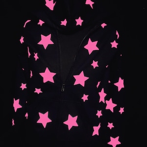 Puede incluir: Una sudadera negra con cremallera y un patrón de estrellas de color rosa brillante distribuidas por la parte delantera y la capucha. La prenda incluye un cierre de cremallera central y mangas largas, con el diseño de estrellas en el torso.