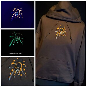 May include: A dark brown hoodie with a black and yellow spider embroidered on the front. The spider glows in the dark, with green and yellow accents.