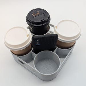 May include: A gray plastic cup holder with six compartments, holding four disposable coffee cups and a black smartphone. One of the cups has a black lid with the word "Chai" written on it.