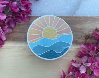 May include: A round sticker with a sunset over the ocean. The sun is yellow with orange and pink rays. The ocean is blue with white waves.