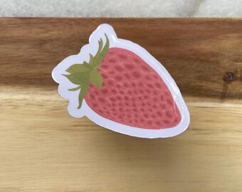May include: A sticker featuring a red strawberry with green leaves. The strawberry has a textured appearance and is outlined in white. The sticker is placed on a wooden surface.