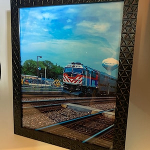Peut inclure: Une photographie encadrée d'un train bleu et rouge avec les mots "Brookfield, IL" sur le côté du train. Le train est sur un ensemble de voies ferrées avec un ciel bleu et des nuages blancs en arrière-plan.