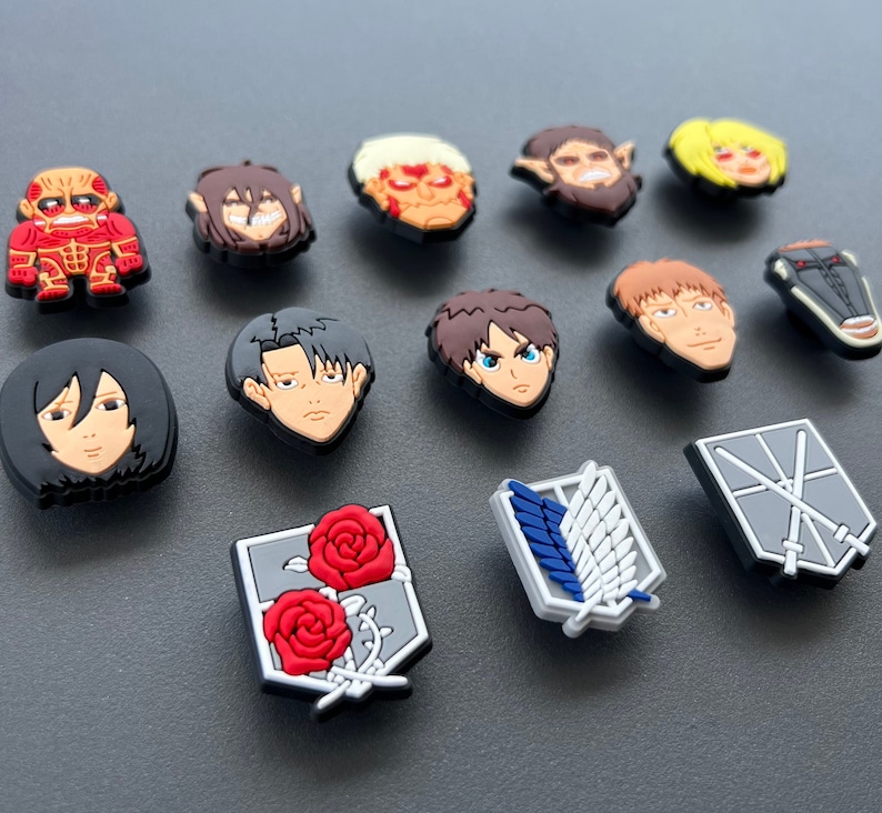 May include: A collection of colorful shoe charms featuring characters and symbols from the anime series Attack on Titan. The charms include character faces and the Survey Corps emblem, in various colors and designs.