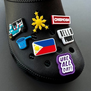 May include: Black shoe with various colorful charms attached. Charms include a blue and white tuk-tuk, a yellow sunburst, a blue and red flag, and text that reads "CHISMOSA", "HELLA PINOY", and "UBE ALL DAY!"
