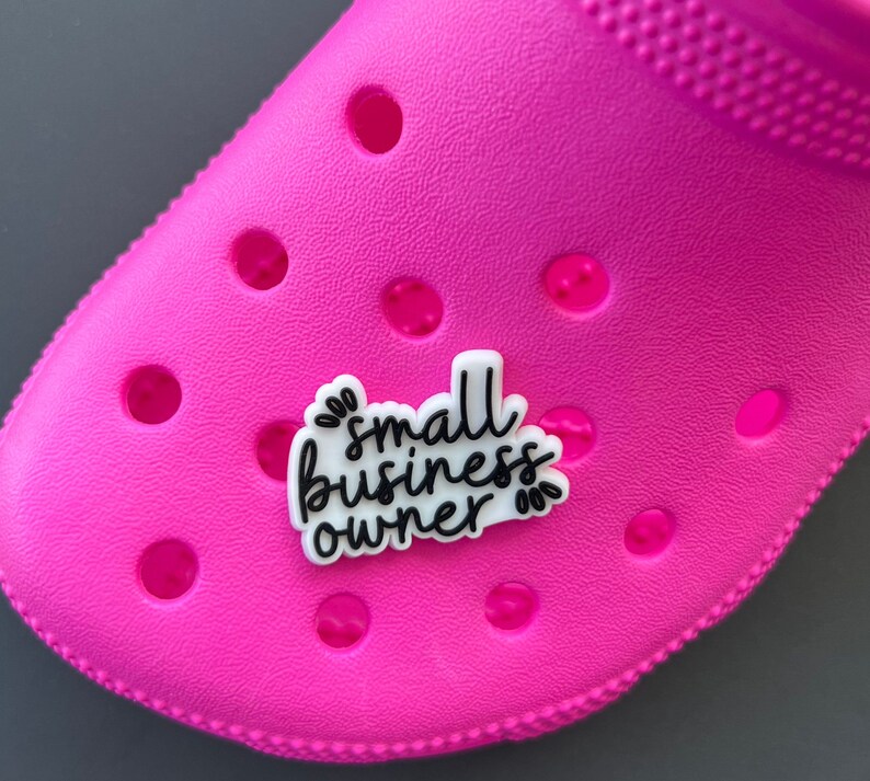 May include: A bright pink shoe with a white and black charm that reads "small business owner" in a handwritten style. The charm is attached to the shoe, which has several round holes.