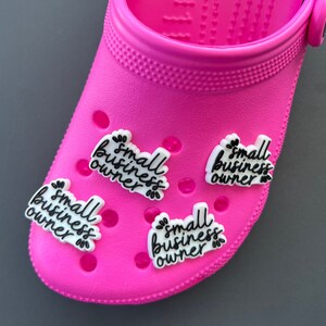 May include: A bright pink clog shoe with several white and black charms. Each charm has the words "small business owner" in a handwritten style font. The charms are decorated with small leaf accents.