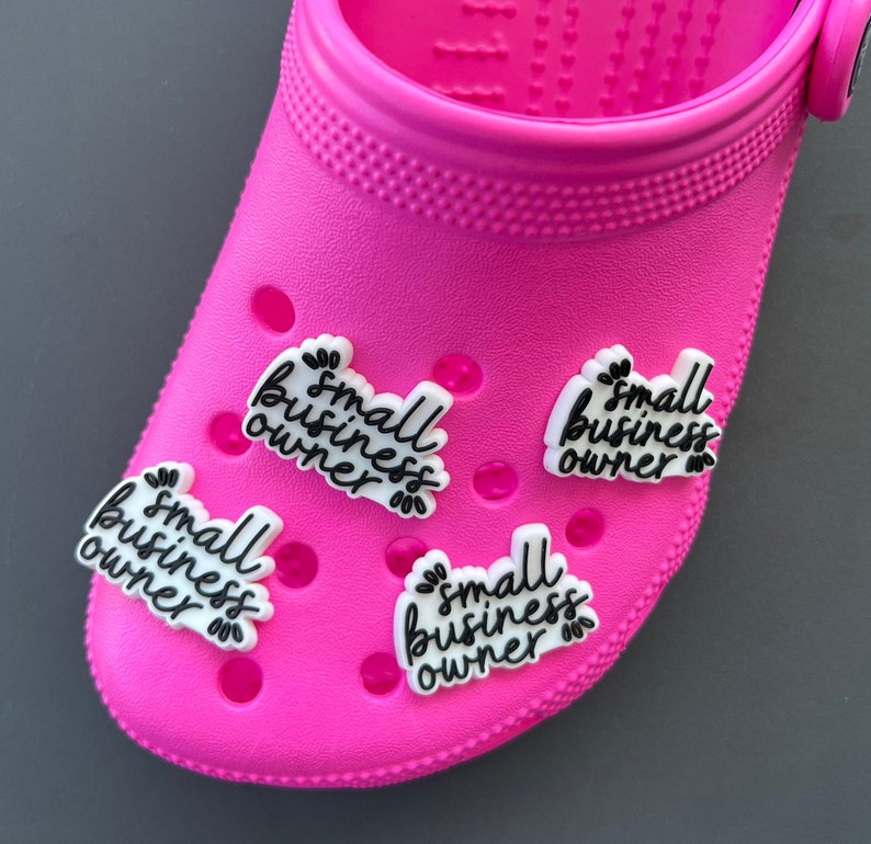 May include: A bright pink clog shoe with several white and black charms. Each charm has the words "small business owner" in a handwritten style font. The charms are decorated with small leaf accents.