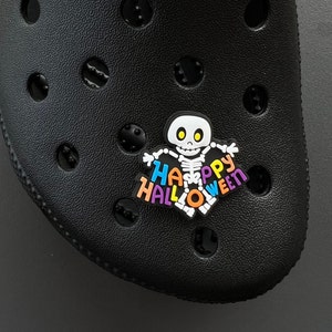 May include: A black shoe with a decorative charm. The charm features a white skeleton with the words "Happy Halloween" in colorful letters. The shoe has multiple holes and a textured surface.