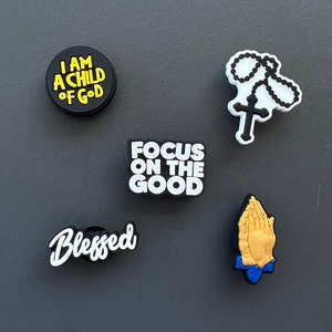 May include: A collection of five black and white shoe charms with religious themes. The charms include the phrases "I AM A CHILD OF GOD", "FOCUS ON THE GOOD", and "Blessed". Other charms depict a rosary and praying hands.