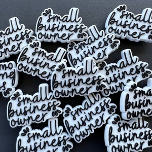 May include: A collection of white and black "small business owner" charms. The charms have a cursive font and decorative floral accents. The charms are scattered on a dark surface.