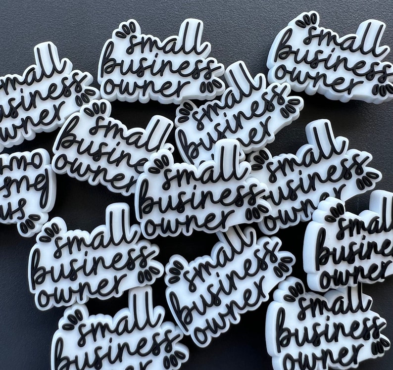 May include: A collection of white and black "small business owner" charms. The charms have a cursive font and decorative floral accents. The charms are scattered on a dark surface.