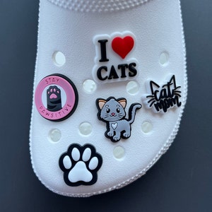 May include: White shoe adorned with various cat-themed charms. Charms include a paw print, a gray cat, the phrase "I ❤️ CATS," and "Cat mom." One charm reads "Stay Pawsitive."