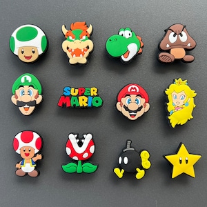May include: A collection of colorful, cartoon character charms, including Mario, Luigi, Princess Peach, and Bowser. Other characters include a mushroom, a Goomba, a Piranha Plant, and a star. The charms are likely for shoes or accessories.