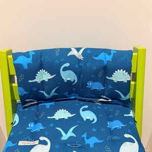 May include: A blue high chair cushion set featuring a repeating pattern of light blue and white dinosaurs, including long-necked sauropods and flying pterosaurs, interspersed with small lightning bolt icons and a small white label reading NiNiLi.
