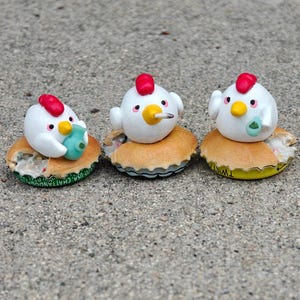 May include: Three whimsical chicken figurines with red combs, yellow beaks, and white bodies, each perched on a bottle cap. One holds a blue object. The bottle caps have a yellow and green design.