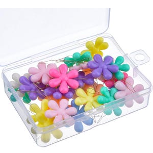 May include: A clear plastic container filled with colorful flower-shaped push pins. The pins are in various colors, including pink, purple, blue, green, and yellow.