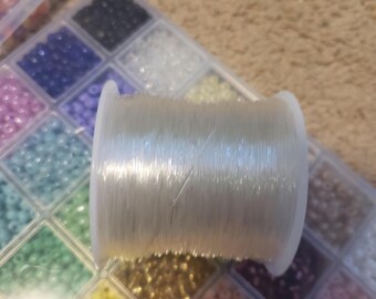 Puede incluir: Un carrete de hilo elástico transparente para hacer joyas, junto a un contenedor de cuentas de colores.