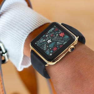 Könnte beinhalten: Eine goldfarbene Smartwatch mit schwarzem Armband wird am Handgelenk getragen. Das Zifferblatt zeigt ein Blumenmuster mit roten, weißen und grünen Akzenten. Die Uhr wird mit einem weißen Pullover und einer braunen Ledertasche kombiniert.