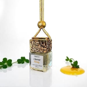 May include: A car air freshener with a gold-coloured decorative top and a clear glass bottle. The label reads "Clover honey" and "Car Diffuser (8ml)". The air freshener is suspended by a gold cord. A small clover and a honey-coloured liquid are also in the image.