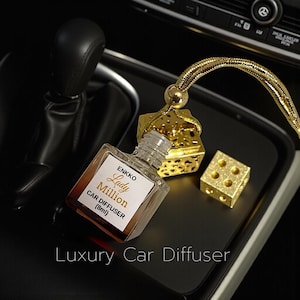 May include: A car air freshener featuring a gold dice design and a small glass bottle labelled "Lady Million Car Diffuser (8ml)". The product is displayed on a black surface, with the text "Luxury Car Diffuser" at the bottom.
