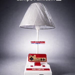 May include: A novelty lamp with a white base and a red and white controller design. The lamp has a grey lampshade and the text "Lampe Famicom". This lamp is a unique piece of home decor, inspired by retro gaming.