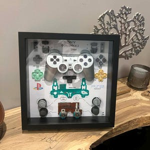 May include: A framed display of a deconstructed Sony DualShock 2 controller. The black frame contains the controller's parts, including buttons, circuit boards, and joysticks, set against a white backdrop. The text "SONY DUALSHOCK 2 2000" is displayed.