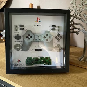 Może przedstawiać: Oprawione dzieło sztuki przedstawiające rozebrany kontroler PlayStation. Obraz zawiera elementy kontrolera, w tym przyciski, pady kierunkowe i płytę główną. Widoczne są logo PlayStation i Sony.