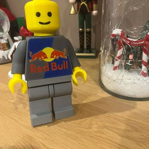 Könnte beinhalten: Eine große Lego-Figur mit gelbem Kopf und Händen sowie grauem Körper. Der Torso der Figur zeigt das Red Bull-Logo in Rot und Blau. Die Figur steht auf einer Holzoberfläche.