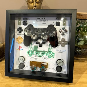 Könnte beinhalten: Ein gerahmter Schattenkasten mit einem zerlegten PlayStation 2 DualShock 2 Controller. Der schwarze Rahmen umgibt die Komponenten des Controllers, einschließlich Tasten, Leiterplatten und dem Controller selbst. Der Text "SONY DUALSHOCK 2 2000" ist sichtbar.