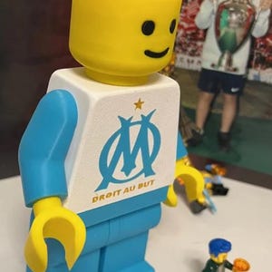 May include: A large, yellow-headed Lego figure with blue arms and legs, and a white torso featuring the Olympique de Marseille logo and the text "DROIT AU BUT". Small Lego figures are in the foreground.