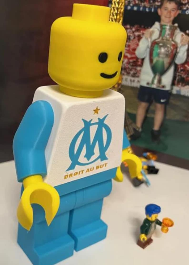 May include: A large, yellow-headed Lego figure with blue arms and legs, and a white torso featuring the Olympique de Marseille logo and the text "DROIT AU BUT". Small Lego figures are in the foreground.