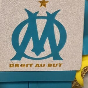 May include: A white and teal emblem with a gold star and the text "DROIT AU BUT". The emblem features a stylized "M" shape in teal. The background is white and teal.