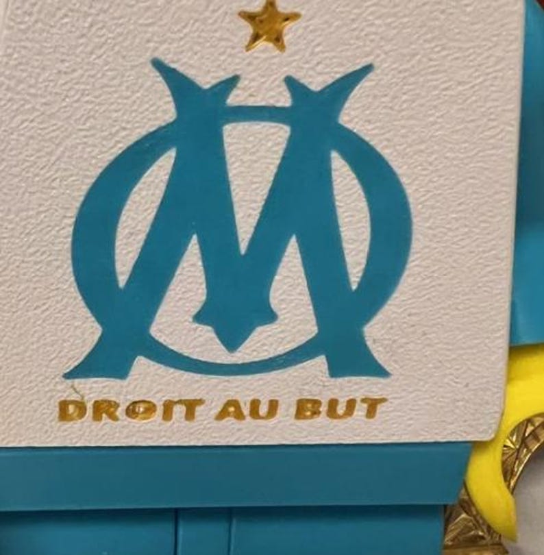 May include: A white and teal emblem with a gold star and the text "DROIT AU BUT". The emblem features a stylized "M" shape in teal. The background is white and teal.