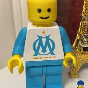 May include: A Lego figure with a yellow head and hands, blue arms and legs, and a white torso. The torso features a blue logo and the text "DROIT AU BUT". A small Lego figure and a gold Eiffel Tower replica are in the background.