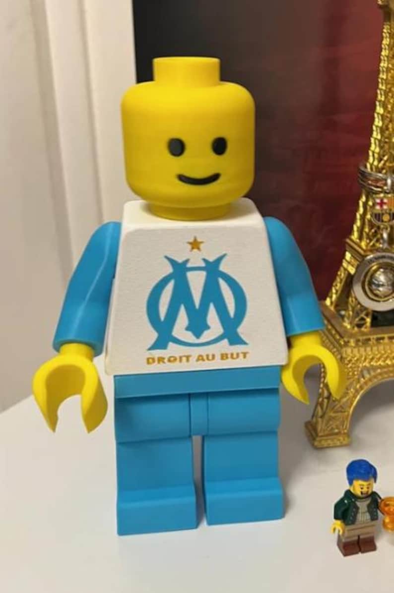 May include: A Lego figure with a yellow head and hands, blue arms and legs, and a white torso. The torso features a blue logo and the text "DROIT AU BUT". A small Lego figure and a gold Eiffel Tower replica are in the background.