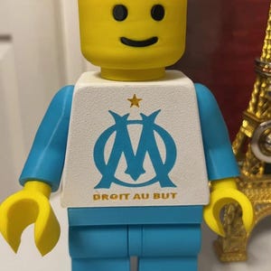 May include: A yellow Lego figure with a smiling face, blue arms and legs, and a white torso. The torso features a blue logo with a star and the text "DROIT AU BUT".