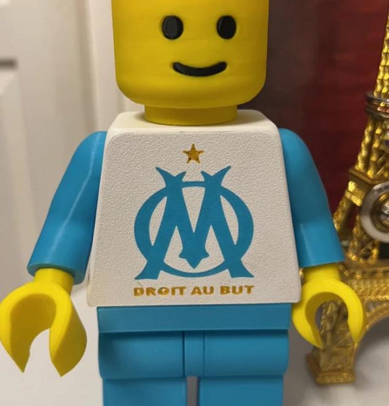 May include: A yellow Lego figure with a smiling face, blue arms and legs, and a white torso. The torso features a blue logo with a star and the text "DROIT AU BUT".