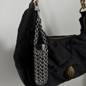 May include: A black quilted nylon shoulder bag with a thick antique gold chain strap and a metallic eagle head emblem. A silver-toned chainmail lighter holder is attached to the bag's side via a small lobster clasp, containing a black lighter.