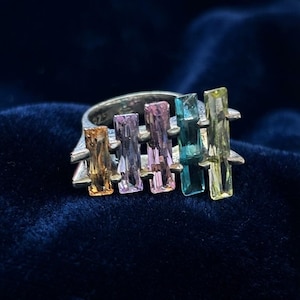 Op de afbeelding: Een zilveren ring met vijf verticaal geplaatste, rechthoekige edelstenen op een rij. De stenen hebben een kleurverloop van oranje, lichtpaars, roze, blauwgroen en geel. De metalen ring is gestempeld met het getal 925.