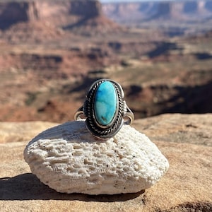 Op de afbeelding: Een zilveren ring met een ovaalvormige turkoois steen met blauwe en groene marmering. De steen is gezet in een gladde zetting, omringd door een gedraaide touwrand en decoratieve zilveren metalen accenten aan de zijkanten van de ring.