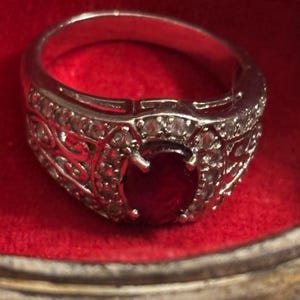 Op de afbeelding: Een zilverkleurige ring met een centrale ovaal geslepen dieprode edelsteen die door vier pootjes wordt vastgehouden. De band is versierd met kleine heldere steentjes en ingewikkelde opengewerkte krulpatronen langs de zijkanten.