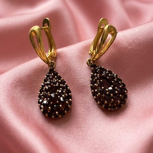 May include: A pair of gold drop earrings featuring teardrop-shaped pendants encrusted with small, dark red garnet gemstones. The earrings have secure lever-back closures and a polished gold finish, showcasing a cluster of faceted stones.