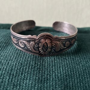 Könnte beinhalten: Ein Armreif aus silberfarbenem Metall mit offenem Design. Die Oberfläche ist mit detaillierten Gravuren in Form von Ranken und floralen Mustern verziert, die durch dunkle, oxidierte Vertiefungen hervorgehoben werden und einen Kontrast bilden.