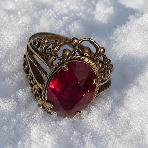 May include: A gold-toned metal ring featuring an oval-shaped, faceted deep red gemstone set in the center. The band and setting are crafted with an intricate, openwork filigree design that includes small spherical accents along the top edge.