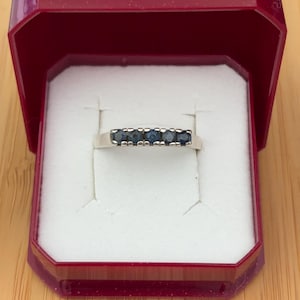 May include: A silver-toned band ring featuring five small, round, dark blue stones set in a row across the top. The ring is presented inside a deep red, open jewellery box with a white interior lining designed to hold the ring securely in place.